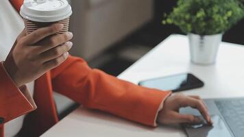 giovane asiatico donna, digitale nomade Lavorando remoto a partire dal un' bar, potabile caffè e utilizzando computer portatile, sorridente. video