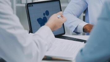 multiraciaal medisch team hebben een vergadering met artsen in wit laboratorium jassen en chirurgisch scrubs gezeten Bij een tafel bespreken een patiënten verslagen, medisch team controle x-ray resultaten. video