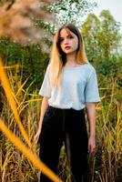 hermosa joven niña vistiendo blanco gris camiseta y negro pantalones posando en contra alto verde y amarillo césped en temprano calentar otoño. al aire libre retrato de hermosa hembra modelo foto