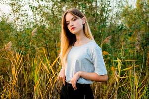 hermosa joven niña vistiendo blanco gris camiseta y negro pantalones posando en contra alto verde y amarillo césped en temprano calentar otoño. al aire libre retrato de hermosa hembra modelo foto