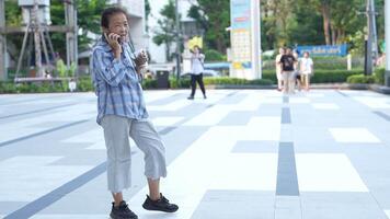 asiático mayor antiguo mujer es Bebiendo café y caminando alrededor al aire libre peatonal. video