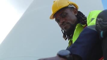 africano hombre trabajadores Ingenieria sentado con confianza con azul trabajando traje vestir y la seguridad casco en frente de viento turbina. concepto de inteligente industria trabajador operando de renovable energía. video