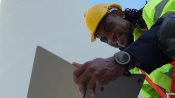africano hombre trabajadores Ingenieria sentado con confianza con azul trabajando traje vestir y la seguridad casco en frente de viento turbina. concepto de inteligente industria trabajador operando de renovable energía. video