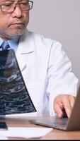 Vertikale, ein Arzt im ein Weiß Labor Mantel ist Sitzung beim ein Schreibtisch mit ein Laptop offen. er ist suchen beim das Bildschirm aufmerksam video
