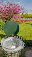 Public trash can with an open lid in a colorful spring park with blooming pink trees and flowers, conceptually related to Earth Day and environmental awareness photo