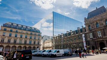 Modern meets classic architecture with a reflective glass building mirroring Pariss Louvre Museum, under a blue sky, photographed on April 14th, 2024 photo