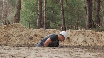 een arbeider vervelend een wit moeilijk hoed is actief graven een versmallen loopgraaf in een bebost Oppervlakte. de omgeving terrein is ongelijk met zichtbaar boom stronken en bodem hopen. video