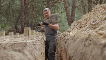 en arbetstagare bär är aktivt grävning en smal dike i en skog område. de omgivande terräng är ojämn med synlig träd stubbar och jord högar. video