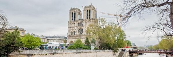 Panoramic view of the iconic Notre Dame Cathedral in Paris on a cloudy day, with surrounding spring foliage and a bustling riverwalk Related to, architecture, travel, heritage photo