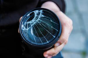 Hands of young photographer holding DSLR photo camera with broken lens filter glass after if fall down onto the floor. Close up. Destroyed cracked photo-filter. Macro. Top view. Selective focus.