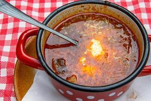 Traditional Slovak Ukrainian Russian cabbage soup with sausage and smoked meat served in retro red metal pot on the plate with spoon. Bowl of delicious Soljanka borsch with white sour cream. Top view photo