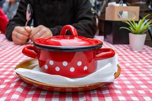 Dining out in the restaurant. Woman is holding the spoon and going to eat the traditional soup in the red cast iron pot served on the ceramic plate with white tissue. Retro style plaid table cloth. photo