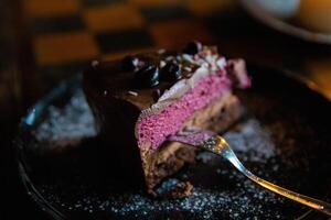 Piece of delicious homemade chocolate cake with blackberries and raspberries dusted with sugar powder served on the dark plate with fork on the wooden chess board table. Tasty Pastry ready for eating. photo