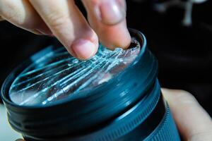 Photographer removing broken pieces of glass out of DSLR photo camera lens filter after if fall down onto the floor. Close up. Destroyed cracked glass of photo-filter. Macro. Top view. Selective focus