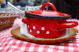 tradicional repollo sopa en el rojo emitir hierro maceta. el cuenco es servido en el cerámico plato con blanco tejido. retro estilo tartán mesa paño. al aire libre restaurante. personas en el borroso antecedentes. foto