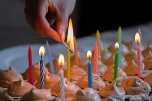 Hand holding matches and lighting colorful candles on the large homemade chocolate birthday cake, decorated with meringues and whipped cream. Close-up. Selective focus. photo
