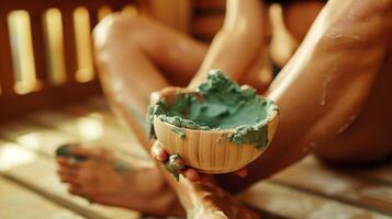 A person sitting crosslegged on a wooden sauna bench with a green herbal paste being applied to their skin by a skilled the. photo