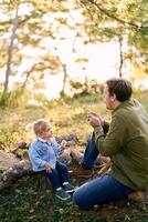 Dad blows on a bouquet of dandelions in his hands on a little girl sitting with her on stumps in the forest photo