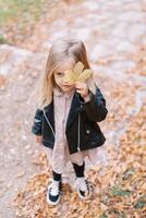 pequeño niña cubre uno ojo con un amarillo hoja mientras en pie en un otoño parque foto