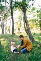 papá sostiene fuera hongos a un pequeño niña sentado en verde césped en el bosque foto