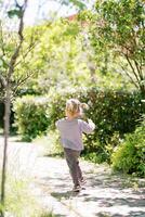 Little girl runs along a path in a green park, waving her arms. Back view photo
