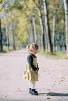 Little girl looks at fallen leaves while standing on the road in an autumn park. Side view photo