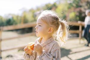 Little girl looks into the distance holding a bitten apple in her hands while standing in the park photo