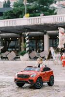 pequeño niña paseos un rojo juguete coche a través de el cuadrado en frente de el edificio foto