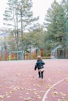 Little girl walks along a sports ground strewn with leaves in an autumn park. Back view photo
