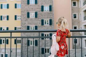 Little girl with a pink toy hare stands by the fence and looks at a colorful residential building. Back view photo