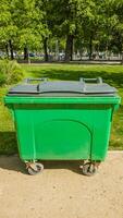 Large green commercial waste bin on wheels in a sunlit urban park setting, highlighting themes of environmental conservation and city sanitation photo