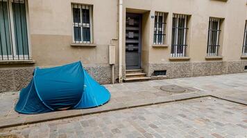 Blue camping tent pitched on a cobblestone urban street, symbolizing homelessness and poverty, with empty European style buildings in the background photo