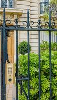 Elegant wrought iron gate with intercom in front of a luxury residential building surrounded by lush greenery, depicting privacy and upscale urban living photo