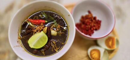 Rawon or Indonesian black beef soup, black color are from indonesian nut called kluwek. Served with lime, chili paste, salted egg, and tempe cracker. Perfect for recipe, article, or any cooking photo