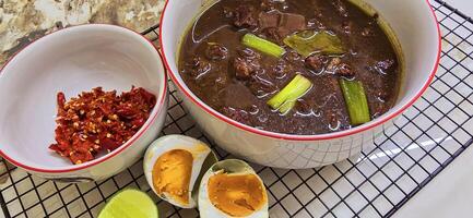 Rawon or Indonesian black beef soup, black color are from indonesian nut called kluwek. Served with lime, chili paste, salted egg, and tempe cracker. Perfect for recipe, article, or any cooking photo