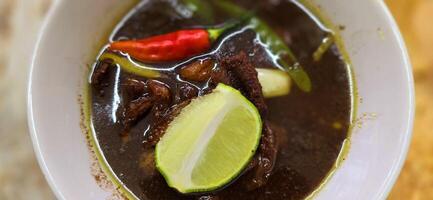 Rawon or Indonesian black beef soup, black color are from indonesian nut called kluwek. Served with lime, chili paste, salted egg, and tempe cracker. Perfect for recipe, article, or any cooking photo