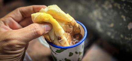 hielo chocolate bebida en un Clásico indonesio hierro jarra con aleatorio verde modelo llamado blirik taza o cangkir blirik en bokeh antecedentes a lo largo con chocolate relleno un pan foto