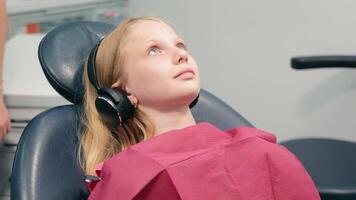 das Mädchen sitzt im das Dental Stuhl und hört zu zu Musik. modern pädiatrisch Zahnheilkunde. video