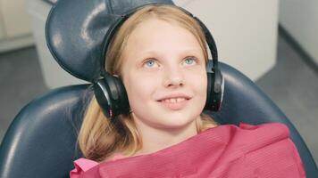 para niños odontología. bebé niña se sienta en el dental silla y sonrisas demostración hermosa dientes. el niña escucha a música y mira a el pantalla video