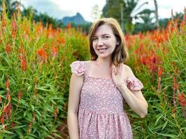 sonriente joven mujer en un floral verano vestir en pie en un vibrante rojo flor campo con un escénico montaña fondo, capturar el esencia de verano foto
