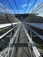 Clear sky over a pedestrian suspension bridge with protective nets, leading into lush greenery. photo