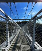 Clear sky over a pedestrian suspension bridge with protective nets, leading into lush greenery. photo