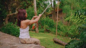 Serene woman sitting outdoors holding a rope, surrounded by lush greenery, embodying tranquility and connection with nature. video