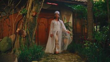 Elegant woman in white dress and headpiece walking through rustic village setting. video