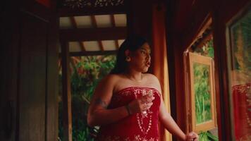 Serene woman in a red dress standing on a porch, gazing out with a thoughtful expression, surrounded by lush greenery. video