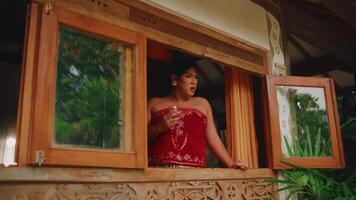 Person in red top leaning out of a wooden window frame, with lush greenery in the background. video