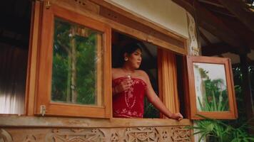 Woman in red dress looking out from a rustic window, with greenery in the background. video
