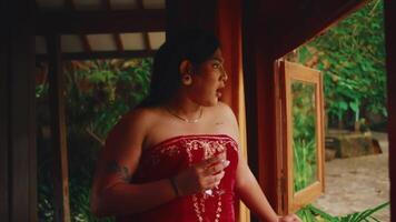 Serene woman in a red dress standing on a porch, gazing out with a thoughtful expression, surrounded by lush greenery. video