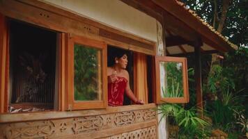 Woman in red dress looking out from a rustic window surrounded by greenery. video