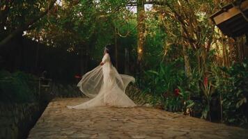 novia en un fluido blanco vestir girando en un jardín camino rodeado por lozano verdor. video